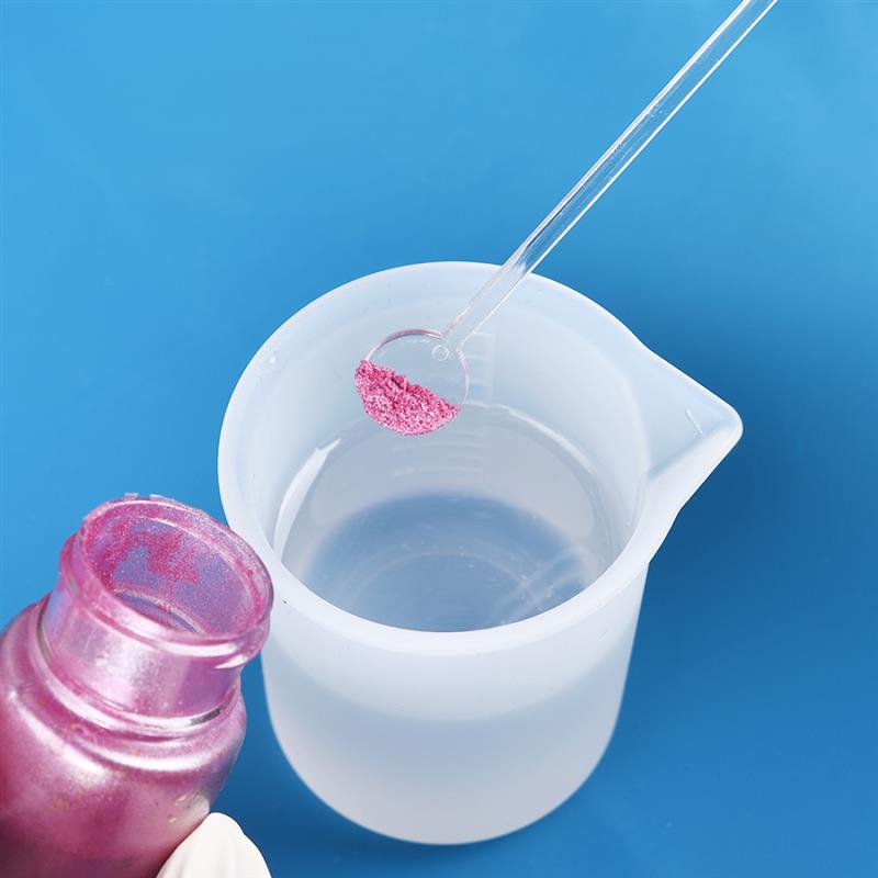 a translucent white plastic measuring cup from an epoxy resin mixing tool set for diy jewelry making holding a clear glass stirring rod with pink glittery powder on the tip