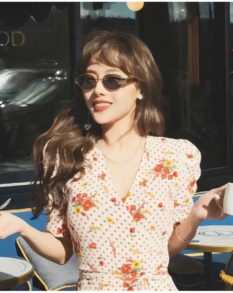 Una mujer que viste un vestido cruzado de color crema con patrones florales rojos y amarillos y pequeños lunares, modela gafas de sol ovaladas para mujer y hombre con montura dorada.