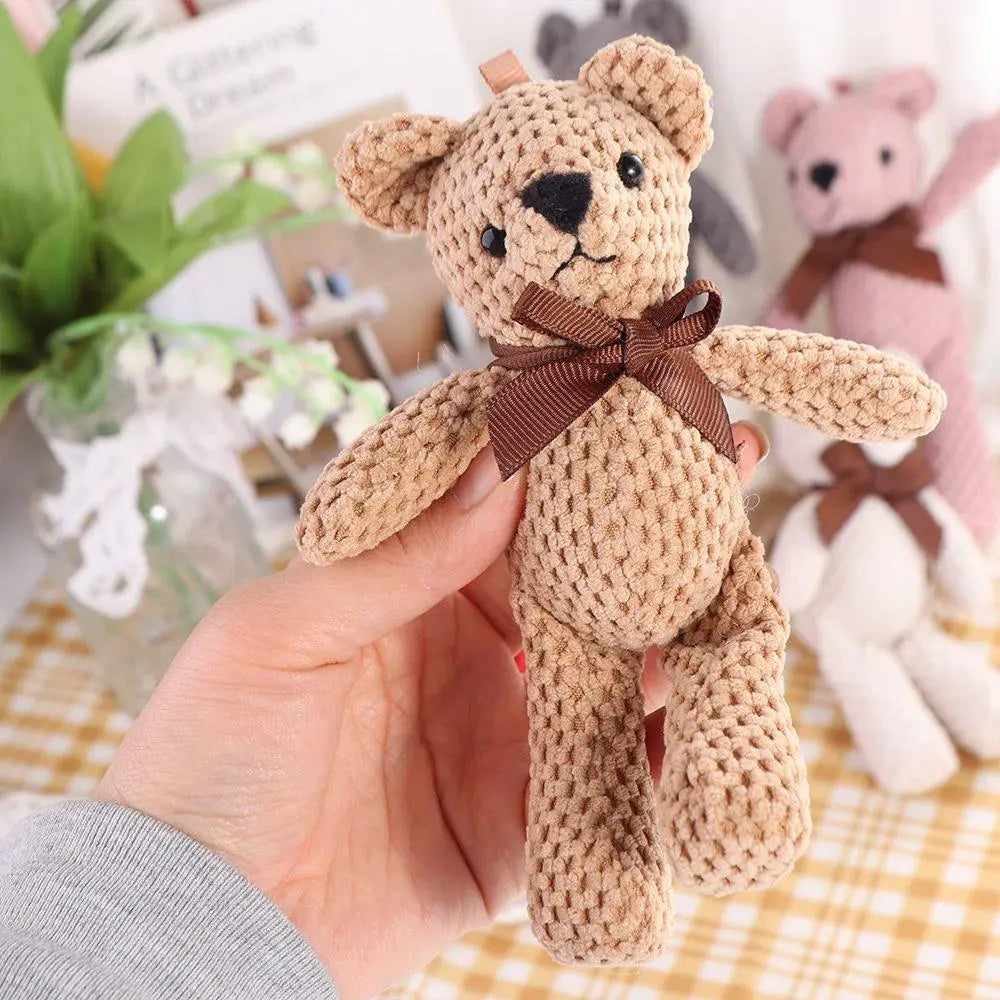 Une peluche d’ourson teddy au crochet beige clair, avec des yeux à boutons noirs, un sourire cousu et un nœud de ruban marron autour du cou.