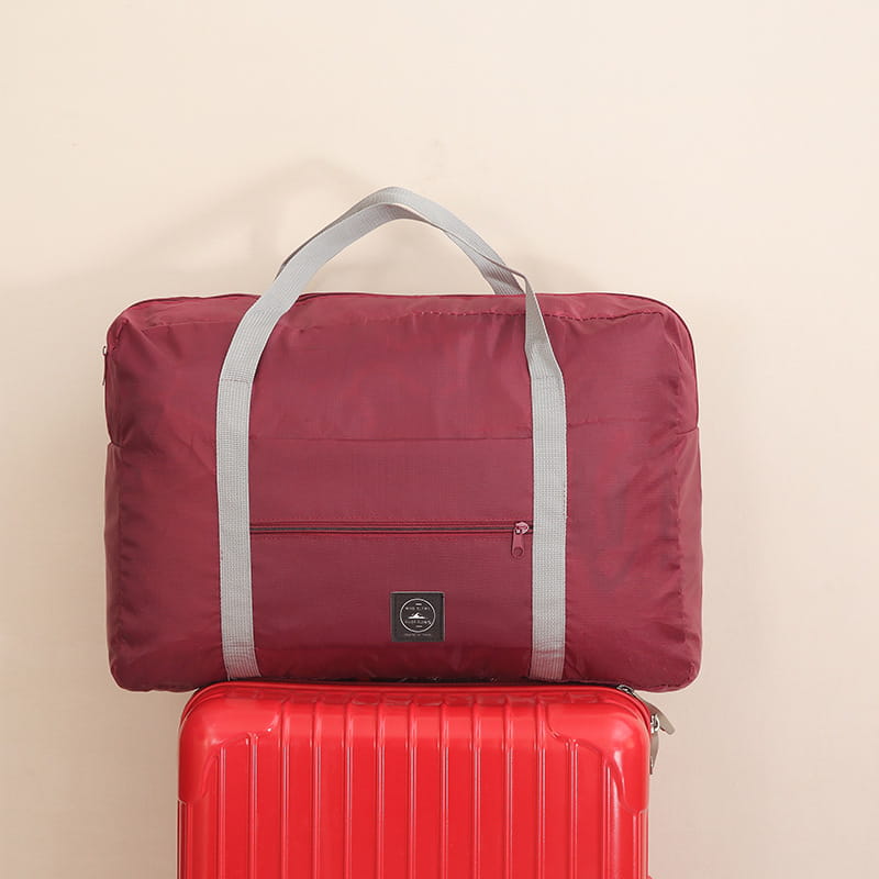 Juego de bolsa de lona de viaje plegable color borgoña con asas de tela grises y un pequeño parche negro con logotipo que se muestra desde la parte frontal.
