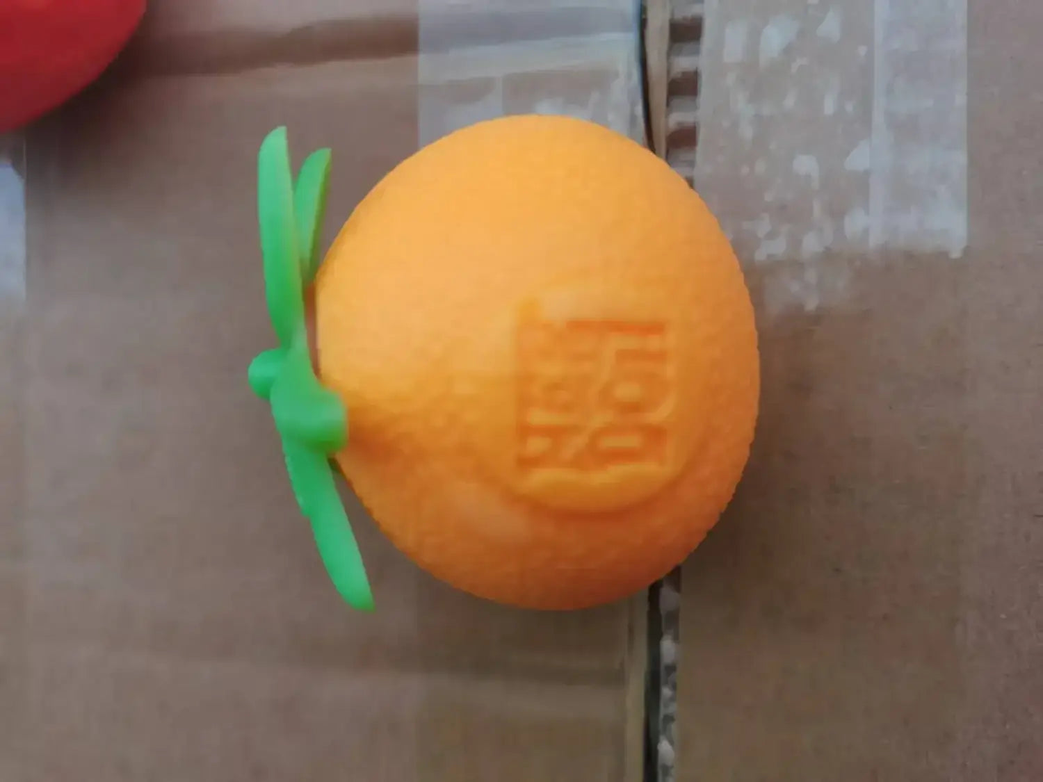 Frontansicht eines orangefarbenen runden quetschbaren Spielzeugs im Erdbeere-Orange-Stressball-Stil mit grünem Kunststoffstiel und eingeprägten chinesischen Schriftzeichen auf der Oberfläche aus dem Erdbeere-Orange-Banane-Stressabbau-Set.