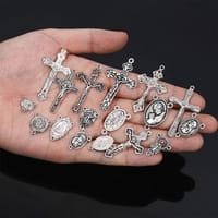 A hand holds a collection of small silver-toned religious pendants and charms including crosses and medallions featured in a Saint Christopher leather keychain antique bronze.