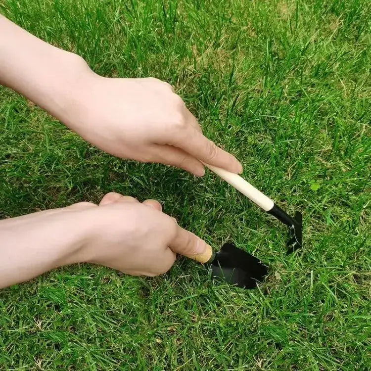 Black metal garden trowel with a light-colored wooden handle from a small gardening tools set being used in grass as a garden shovel.