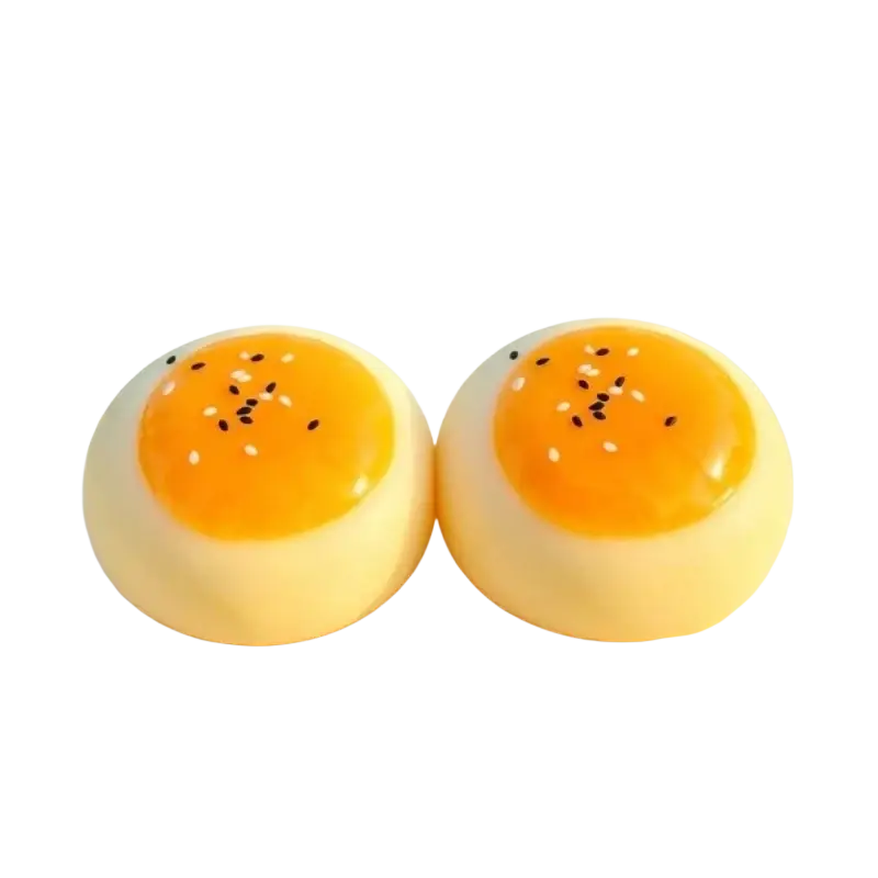 Zwei blassgelbe Eigelb-Gebäckstücke mit glatten Außenflächen und glänzenden orangefarbenen Zentren, die mit schwarzen und weißen Sesamsamen belegt sind, von vorn betrachtet; es sind Silikon-Eigelb-Gebäckstücke, langsam aufsteigend, als knetbare Spielzeuge.