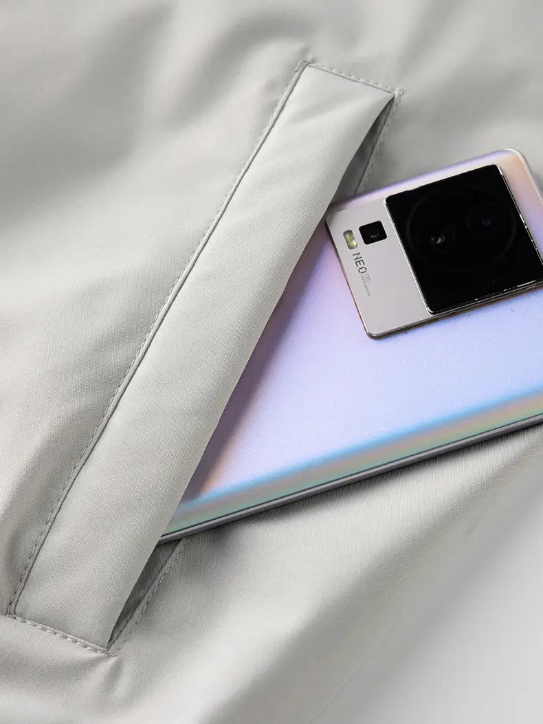 a sleek silver xiaomi 12 pro smartphone with a gradient finish and prominent camera module resting on a light gray fabric surface alongside a windbreaker waterproof hiking jacket for men.