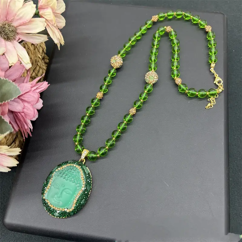 Un collier de pendentif Bouddha vert jade, avec une chaîne à perles facettées en cristal ornée de strass et des accents ton or, adapté à divers âges applicables et doté d’un fermoir à pince en forme de sésame.