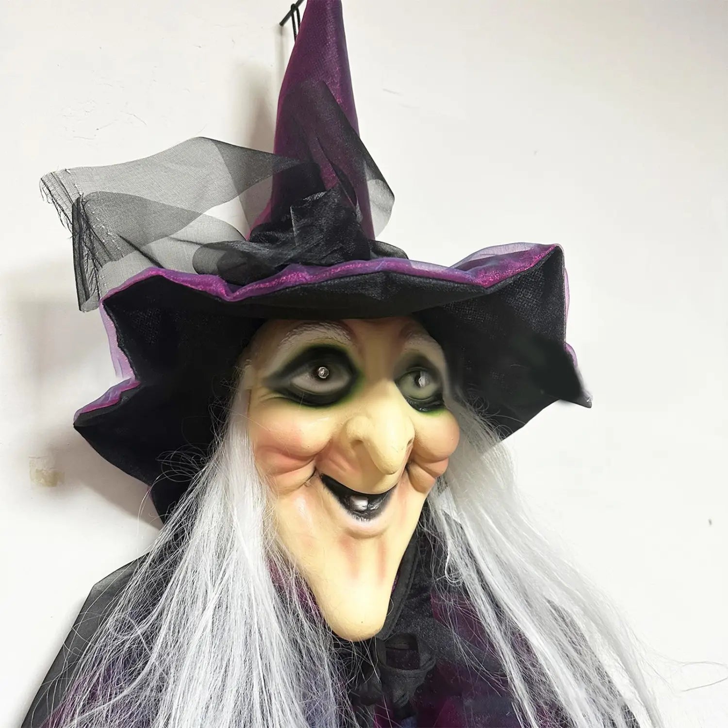 fantôme suspendu de sorcière d’Halloween à activation sonore, avec un chapeau pointu en tissu violet et noir, un visage souriant et de longs cheveux blancs.