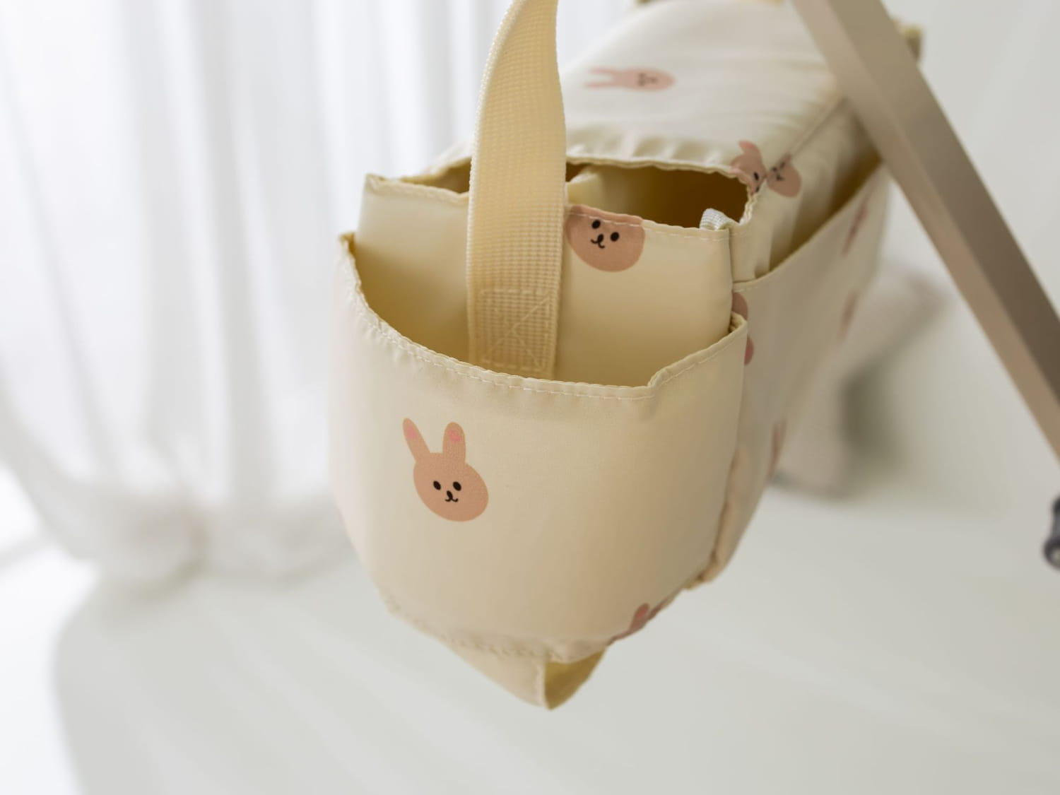 ベージュの布製ベビーカー用オーガナイザーバッグ。クマのデザインで、ピンクのうさぎの顔プリントが入ったベビーボトルキャリア付き。取っ手に合う仕様。