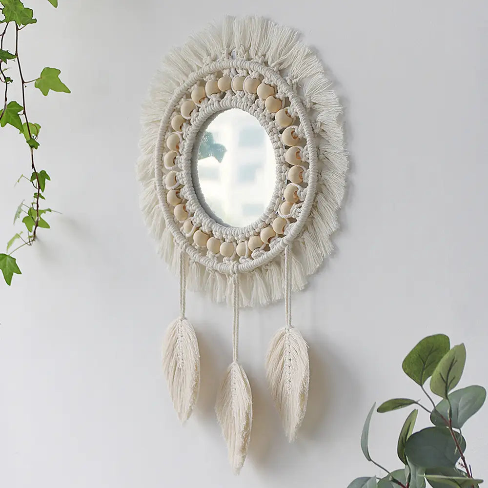 Cream-colored round macrame wall mirror features a circular frame adorned with woven beads, tassels, and three hanging feather-like fringe elements, serving as a decorative mirror for boho interior decor.