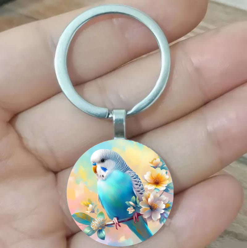 A circular lovebird keychain glass dome bird keyring featuring a vibrant blue and white parakeet perched on a branch with soft yellow and pink flowers set against a metallic silver ring.