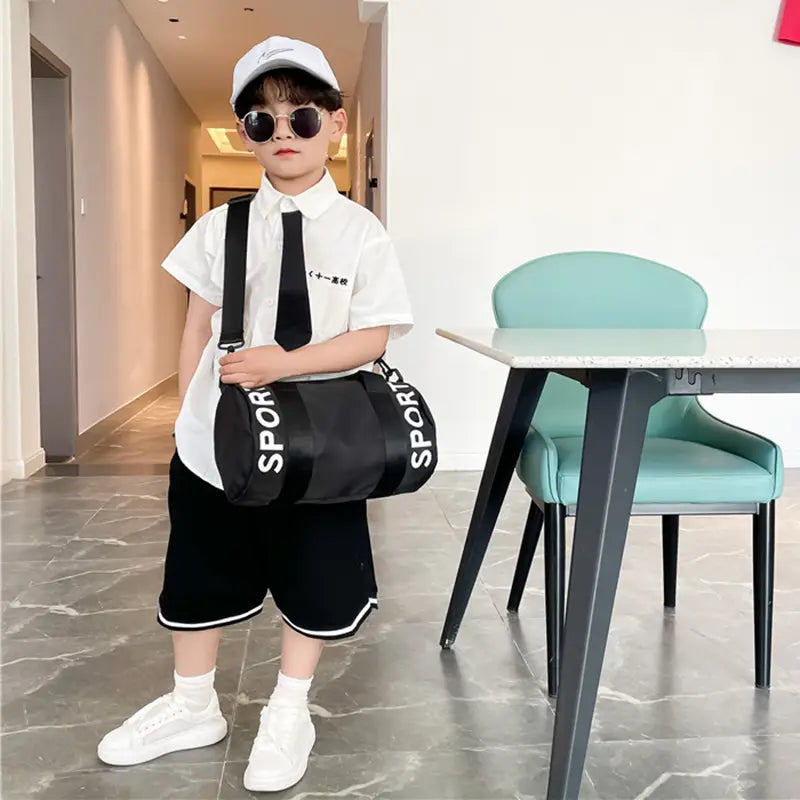Schwarze zylindrische Kinder-Sport-Reise-Sporttasche mit weißer Sportbeschriftung und schwarzen Schulterriemen, entworfen für die Nutzung als Reise-, Gym- und Crossbody-Gepäck.