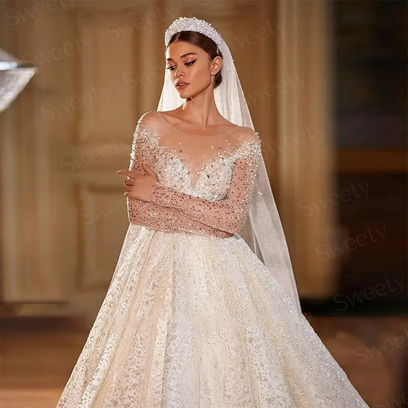 Une robe de mariée en style boule ivoire étincelante, perlée, avec des manches longues épaules dénudées et une jupe en dentelle volumineuse.
