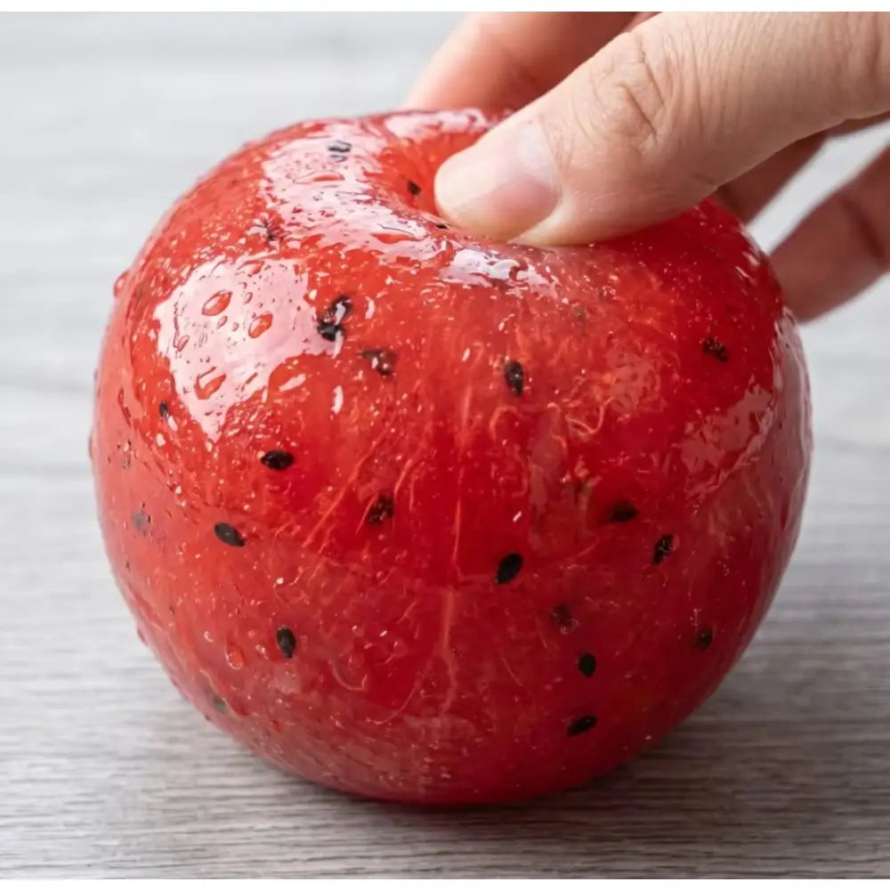 una bola antiestrés artesanal roja brillante con forma de sandía de rebote lento, con motitas negras tipo semillas y gotas de agua, diseñada para aliviar el estrés como juguete de granizado de sandía