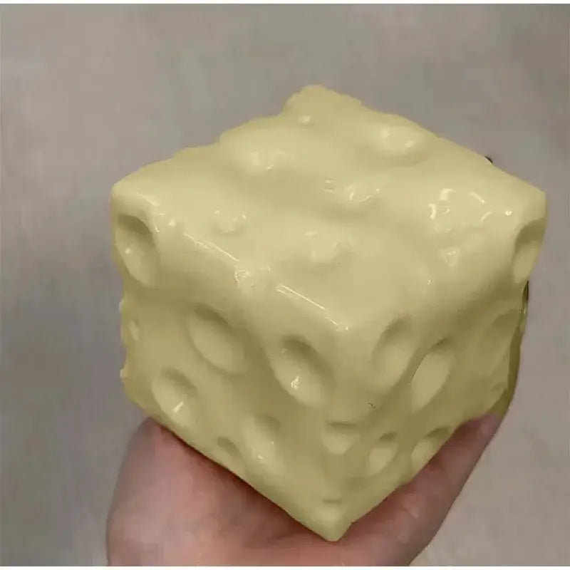 Un juguete antiestrés esponjoso con forma de cubo de queso, de color amarillo pálido y acabado brillante, con una superficie texturizada que imita los orificios del queso suizo.