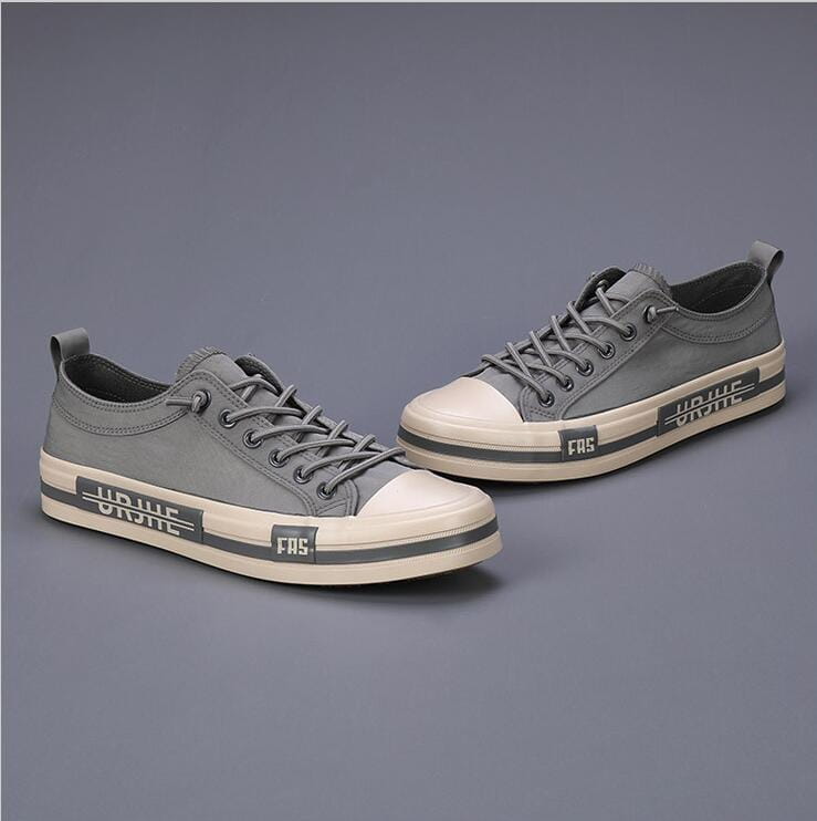 Baskets décontractées en toile pour hommes en gris, avec des coques en caoutchouc blanc pour le bout et des semelles, présentant des rayures noires de branding conçues comme des chaussures d’été légères.