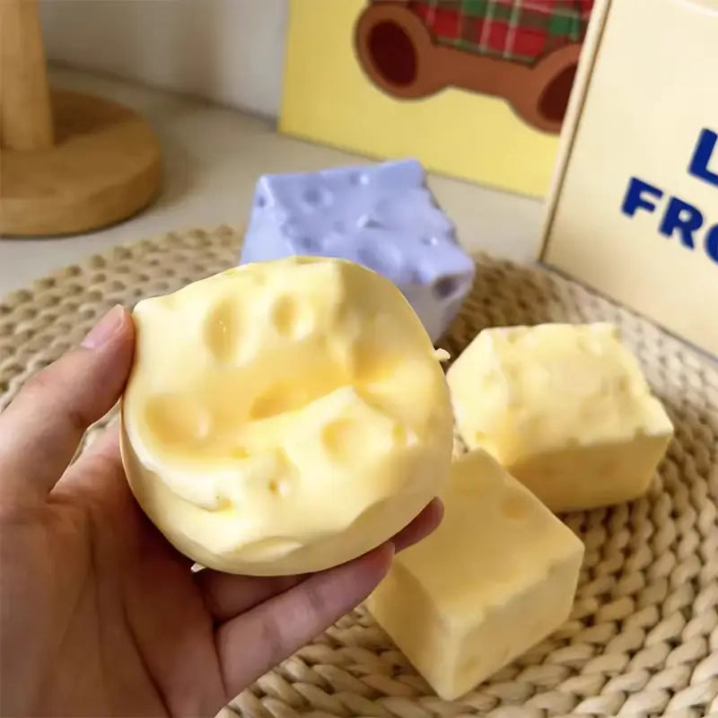 un juguete blando de descompresión de crecimiento lento con forma de queso de color amarillo pálido y aspecto suave, con superficie texturizada que imita los orificios del queso suizo, visto desde el frente.
