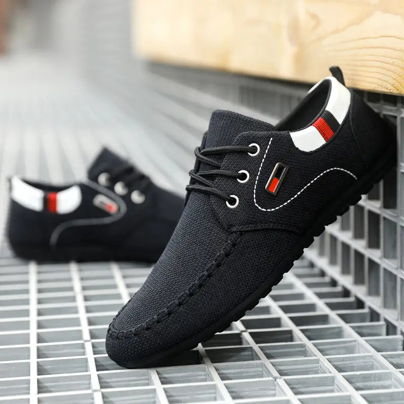 Men’s casual canvas sneakers lightweight daily footwear showing the back side of a black canvas lace-up shoe with white stitching red and black striped accents on the heel tab and a small rectangular logo patch.
