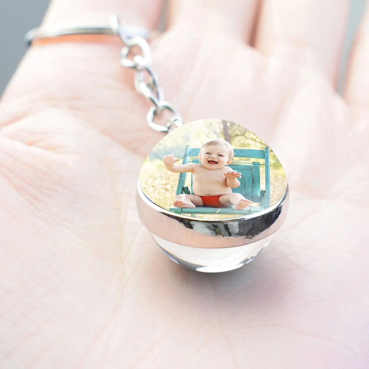 Klarer, runder Glühfoto-Schlüsselanhänger aus Glas mit silberfarbenem Metallrand und Kette, mit einem integrierten Foto eines lächelnden Babys in roten Shorts, das auf einem blauen Stuhl sitzt.