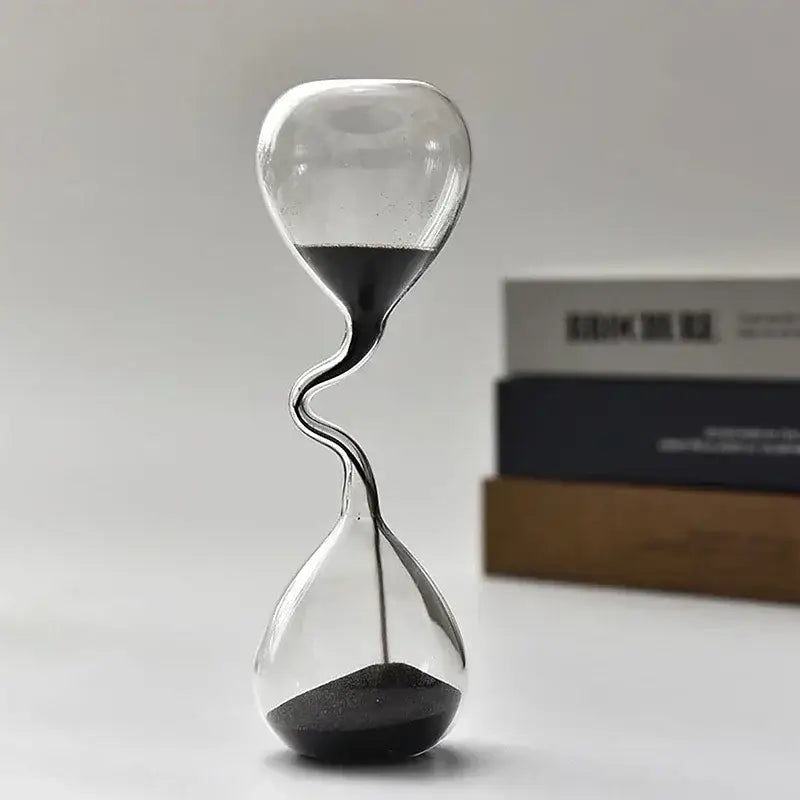 A sleek transparent glass sand timer hourglass with a twisted neck and black sand flowing between two bulbous chambers used as home decoration.