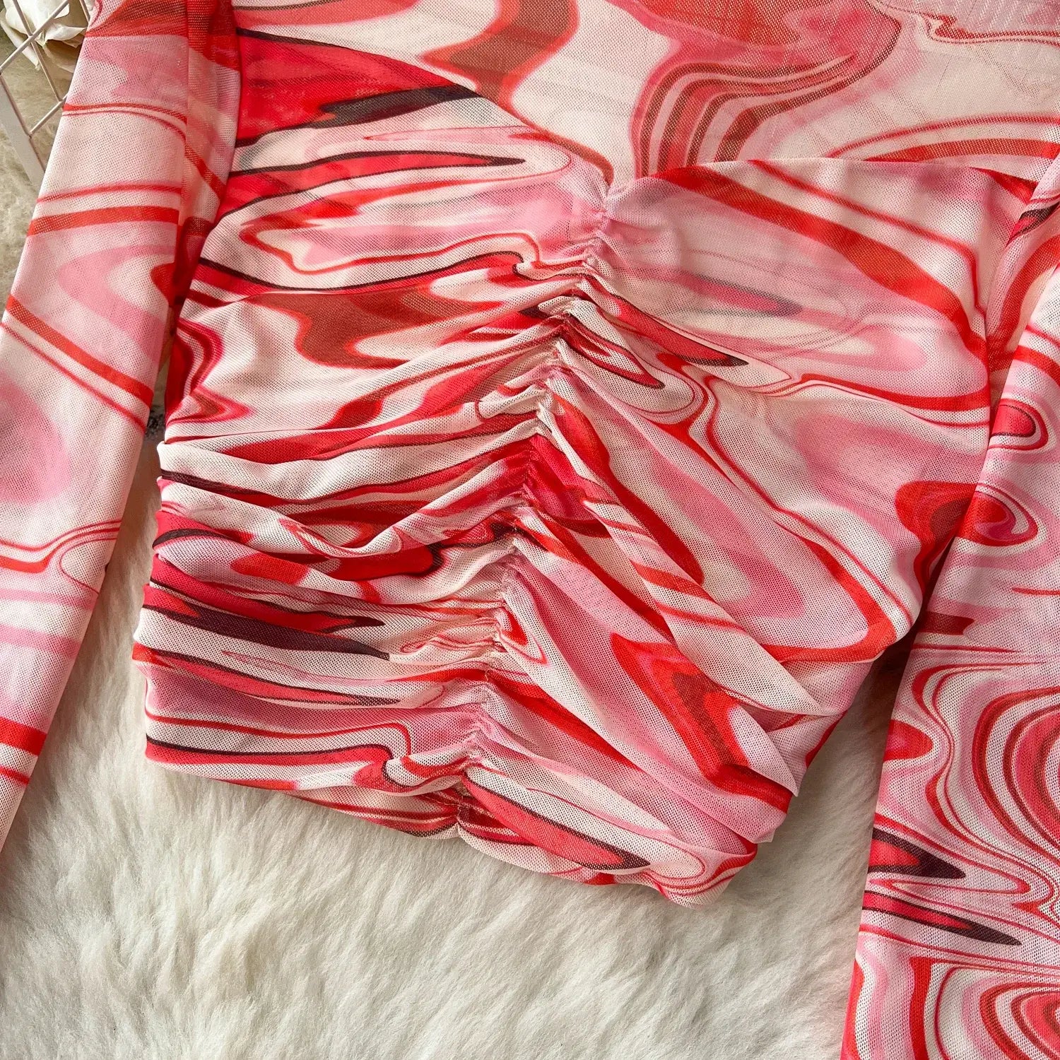 A long-sleeved top from a two-piece mesh skirt set with a vibrant red and pink marbled pattern and ruched detailing at the waist featuring an elastic closure type.
