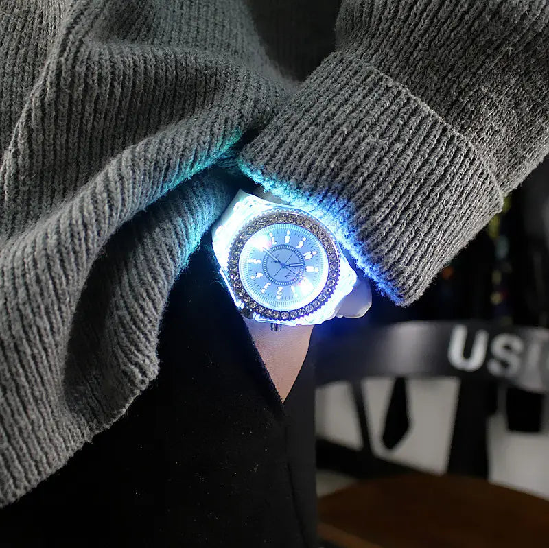 A glowing blue led luminous silicone watch with a white case and rhinestone-adorned bezel is worn on a wrist beneath a gray ribbed sweater cuff.
