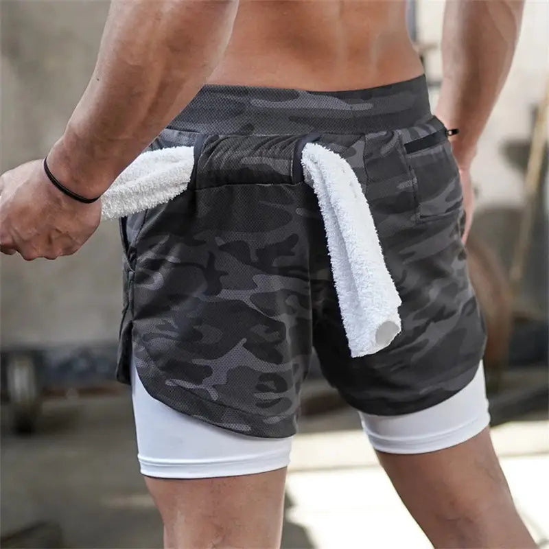 Men 2 in 1 gym shorts with quick dry black and gray camouflage pattern featuring a white inner layer and side pockets holding a rolled up towel for workout activewear.