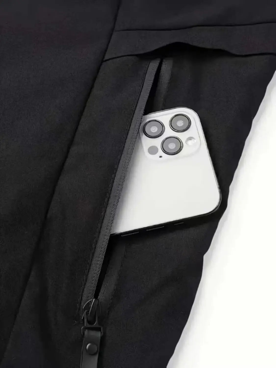 A sleek white iPhone with a triple-camera system resting in the zipper pocket of black fabric pants worn with a men off road hooded jacket windproof casual outerwear.