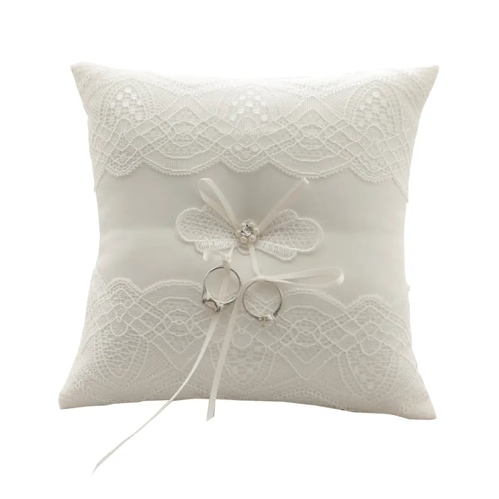Almohada blanca para anillos de boda de encaje con almohadilla portadora de arras que presenta una delicada aplicación de mariposa y dos anillos plateados colocados en su centro.