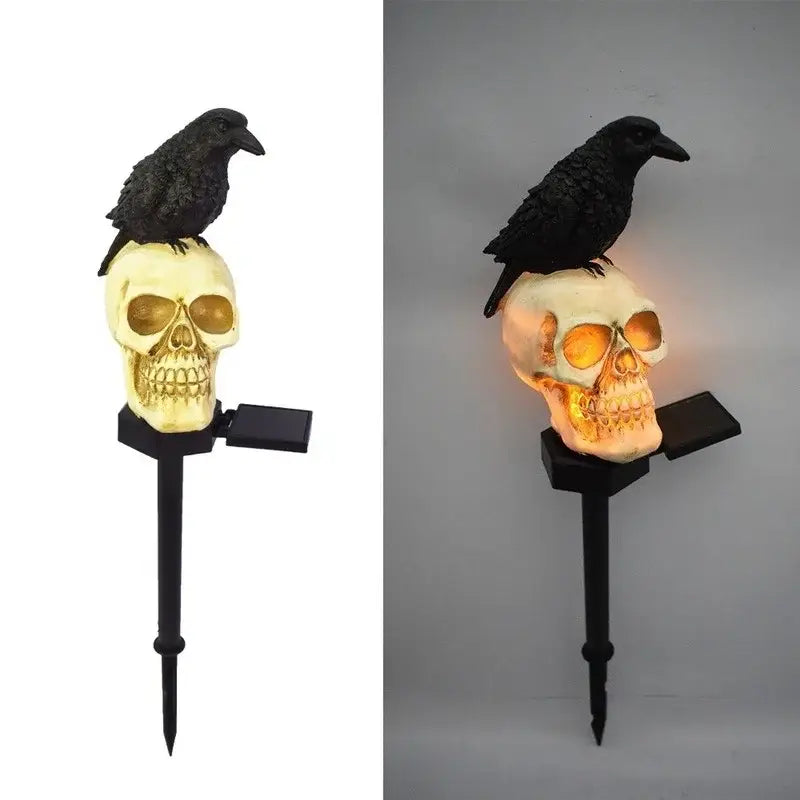 La lámpara de jardín solar de cráneo y cuervo presenta un cuervo negro posado sobre la parte superior de un cráneo color crema con cuencas de los ojos con matices dorados, iluminado mediante una fuente de luz interna.