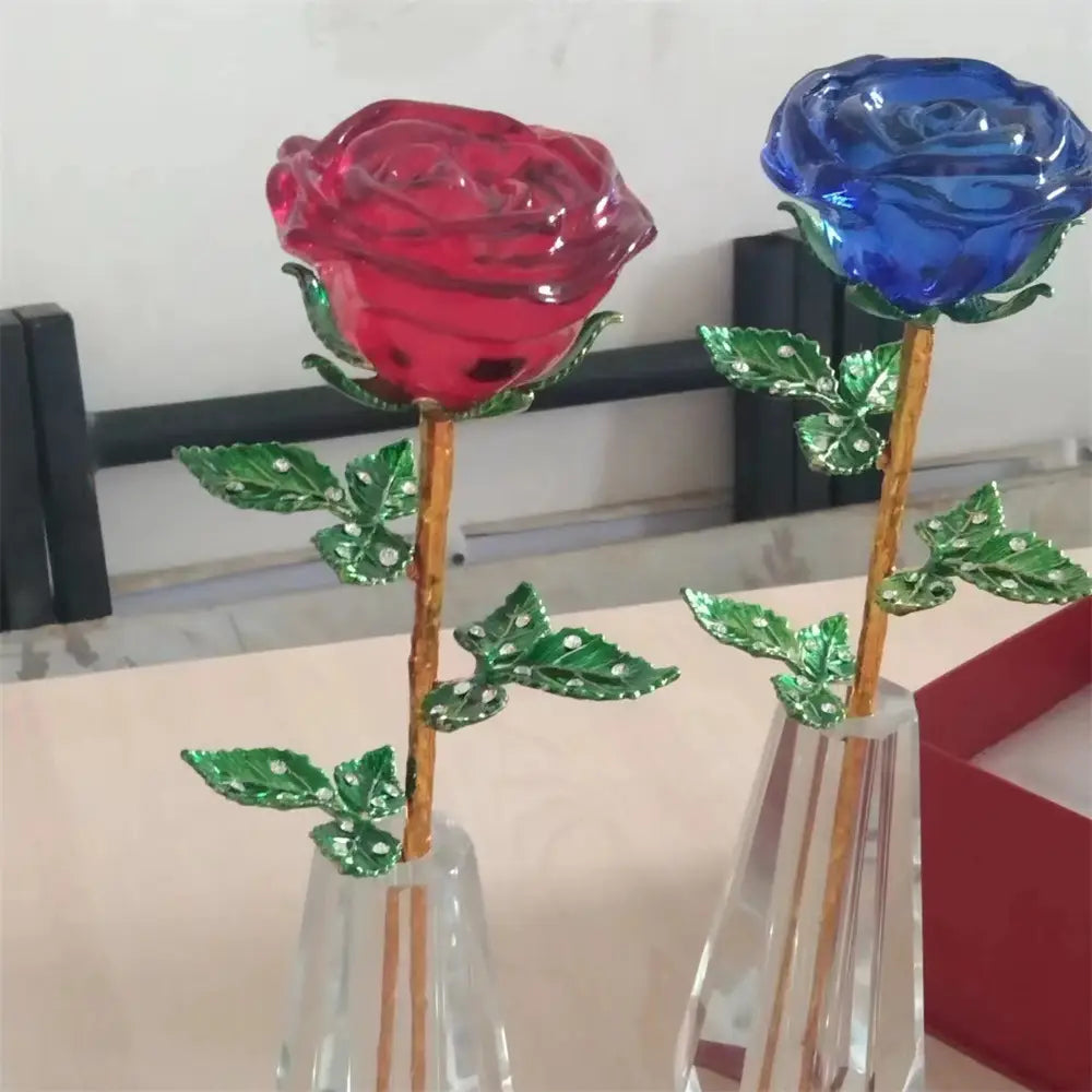 Two translucent crystal glass roses in vibrant red and deep blue with green leaves adorned with tiny white rhinestones and gold-toned stems displayed in clear crystal vases as a Valentine’s Day gift.