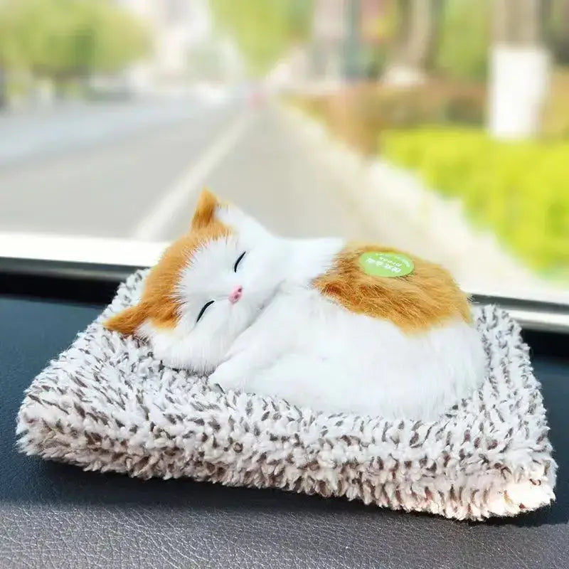 A simulated sleeping cat plush toy with sound featuring a calico design in white and orange fur resting on a textured gray and white fuzzy cushion.