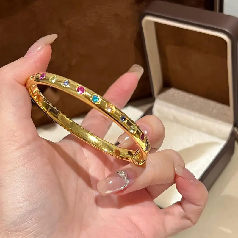Ein Armreifarmband aus goldfarbenem Multigemsteinschmuck mit multicolourfarbenen Edelsteinen, der eine goldfarbene Bandfassung zeigt, die mit einer lebendigen Vielfalt an farbigen Edelsteinen in einem eleganten, durchgehenden Muster verziert ist.