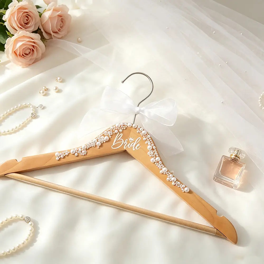 Un cintre de mariage en bois clair pour la mariée, orné de perles blanches et d’une délicate inscription de la mariée, rehaussée d’un nœud en tulle blanc, provenant de l’ensemble de cintres en bois de perle pour la mariée et le marié.