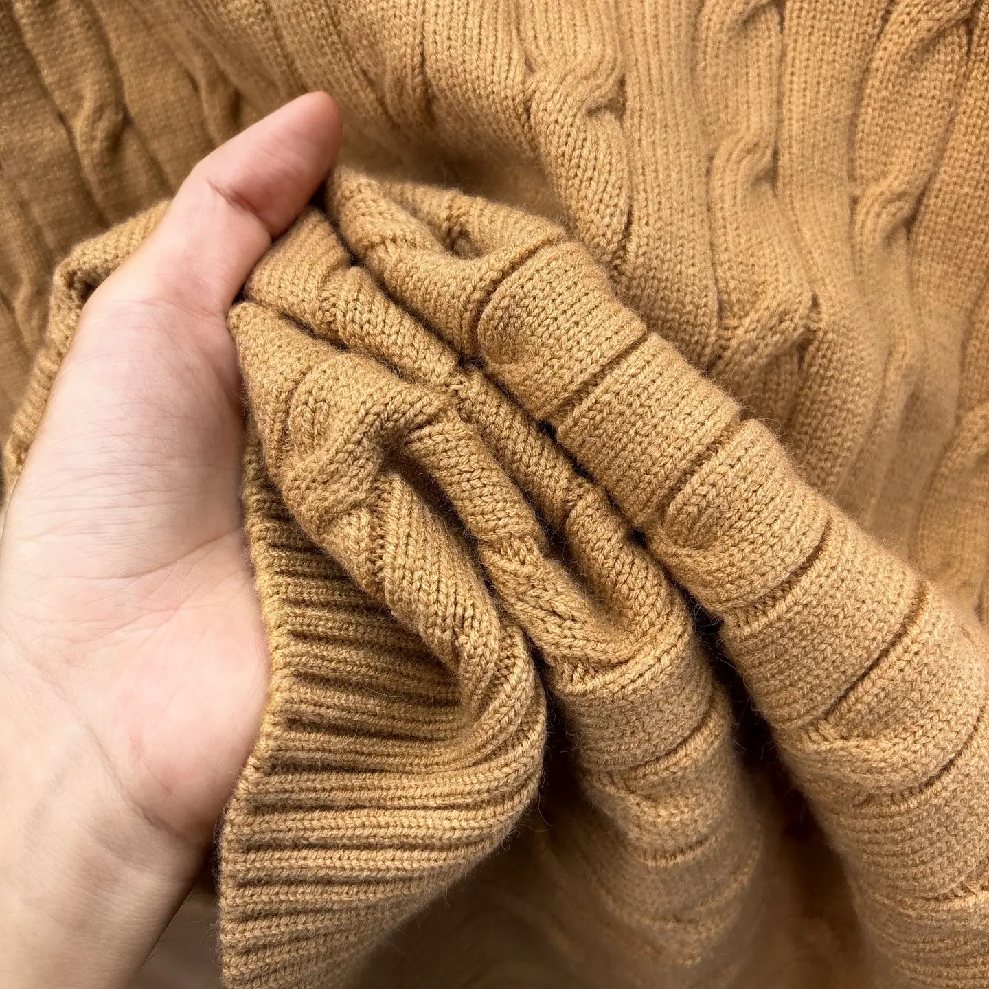 Eine Hand hält einen weichen, sandfarbenen Herren-Designer-Sweater aus Zopfmusterstrick mit halbem Reißverschluss, mit detailreichen strukturierten Mustern.