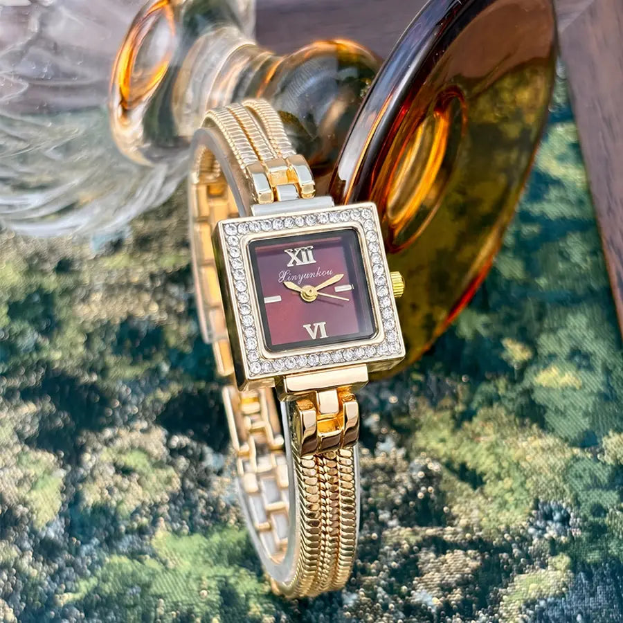 Gold diamond square watch with a gold-toned case, burgundy face featuring roman numerals at xii and vi, and a bezel encrusted with clear crystals on a vintage fringe strap.
