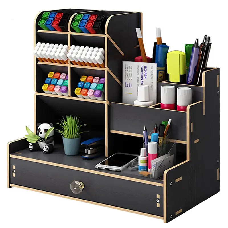 Wooden desk organizer with drawer featuring black and light wood-grain compartments holding markers pens a small plant and other office supplies.
