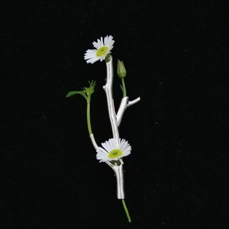 Eine silberfarbene Broschennadel-Blume mit zarten weißen Gänseblümchenblütenblättern, gelben Zentren, grünen Blättern und Knospen an einem verzweigten Stiel, gestaltet als kreatives Corsage-Zubehör.