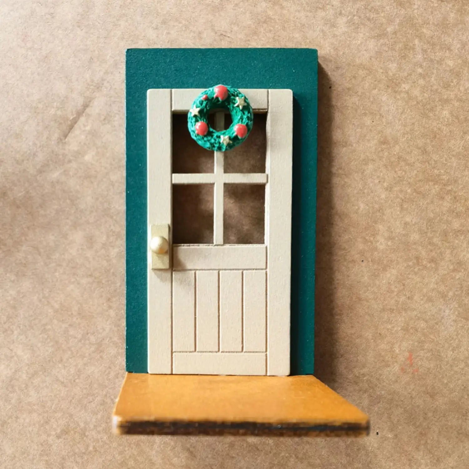 Ornement mural de porte de Noël miniature fabriqué en bois blanc, avec un cadre turquoise et une couronne de fête verte décorée de baies rouges et d’étoiles argentées.