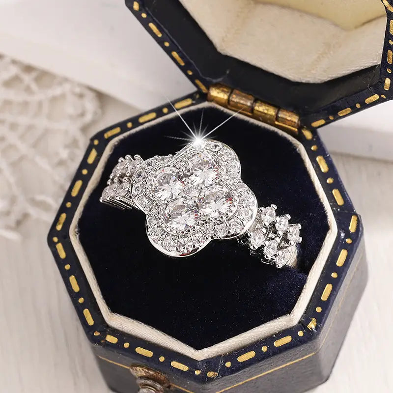 Un anillo de compromiso de trébol de cuatro hojas de plata con brillo, adornado con piedras claras de circonita cúbica, presentado con elegancia en una caja de joyería hexagonal de color negro y dorado.