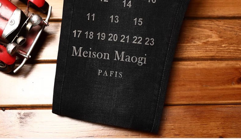 mens slim fit printed black denim jeans shown alongside a black leather-bound calendar with silver embossed text reading meison maogi and paris featuring a grid of dates on its cover.