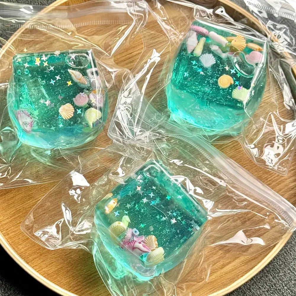 Drei durchscheinende türkisfarbene Resin-Fidget-Toy-Würfel, gefüllt mit Glitzer und miniaturisierten Muscheln, verwendet als ozeanmuschelartige weiche Stressbälle.