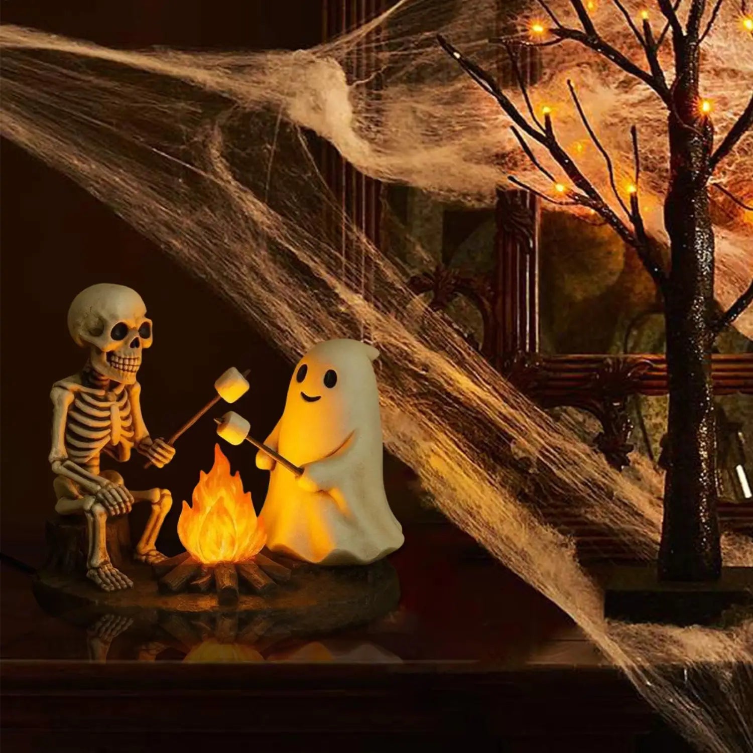 une lanterne de nuit de feu de camp fantomatique blanche et lumineuse avec un visage souriant, assise à côté d’un fantôme squelette et faisant rôtir des guimauves au-dessus d’un feu de camp illuminé, pour la décoration d’Halloween.