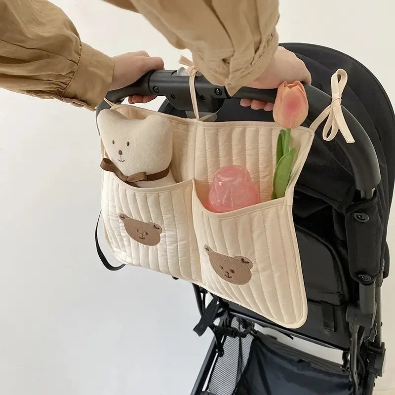 クマの刺繍が施されたクリーム色のキルティング製ベビーカー用オーガナイザー袋で、刺繍のクマの顔と、片方のポケットに取り付けられたふわふわのテディベアが特徴です。