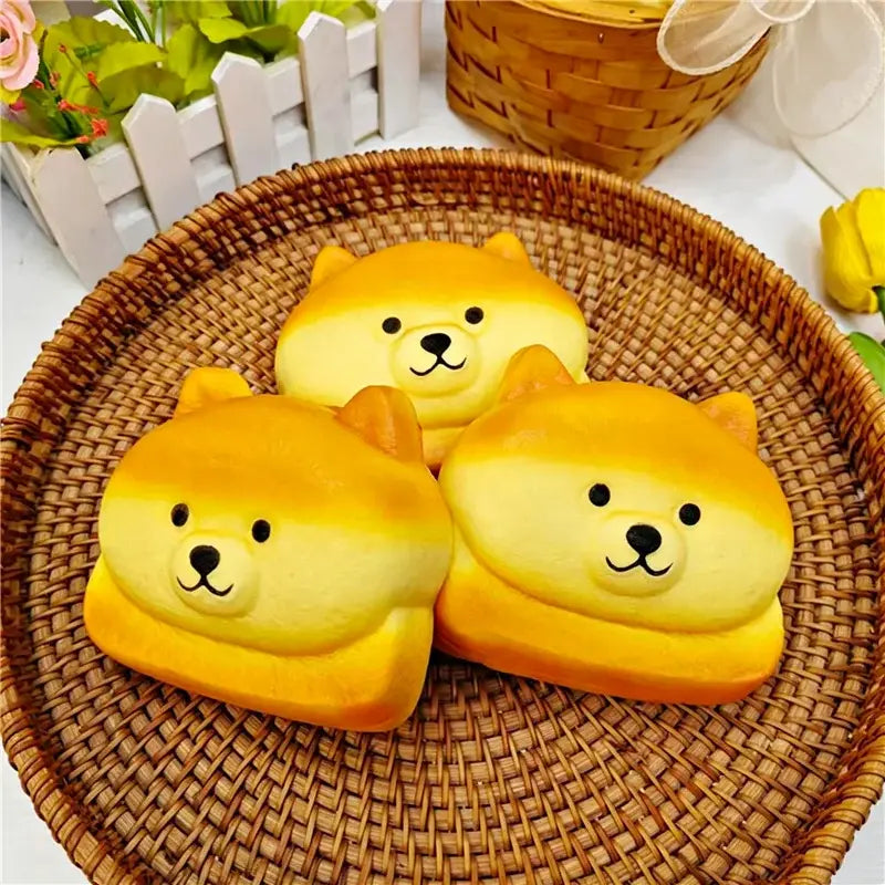 Drei goldgelbe Shiba-inu-ähnliche, langsam aufsteigende, weiche Knetspielzeuge mit schwarzen Augen und Nasen aus Plüschmaterial sitzen in dem Produktbild zum Hundebrot – langsam aufsteigendem, weichem Knetspielzeug – in einem geflochtenen braunen Korb