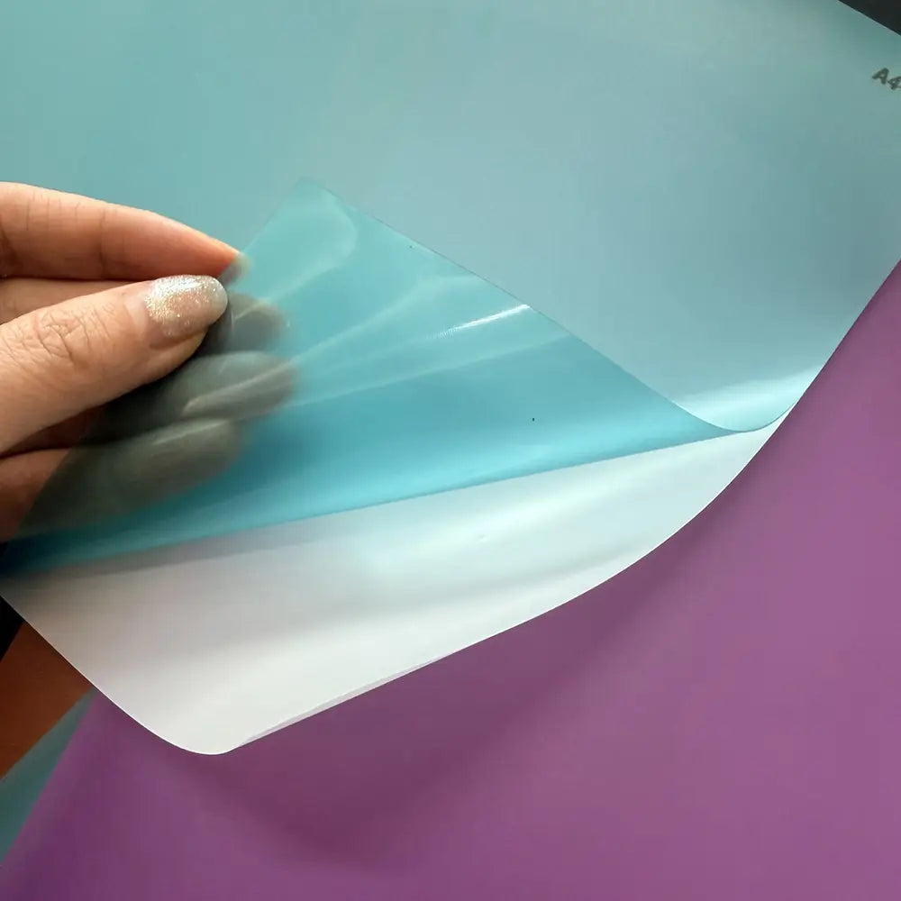 Hoja protectora translúcida azul claro de tamaño A4 con acabado brillante, sostenida por los dedos, para su uso con una máquina de laminación al vacío de película para teléfonos móviles, con el fin de reducir daños por el costo de envío.