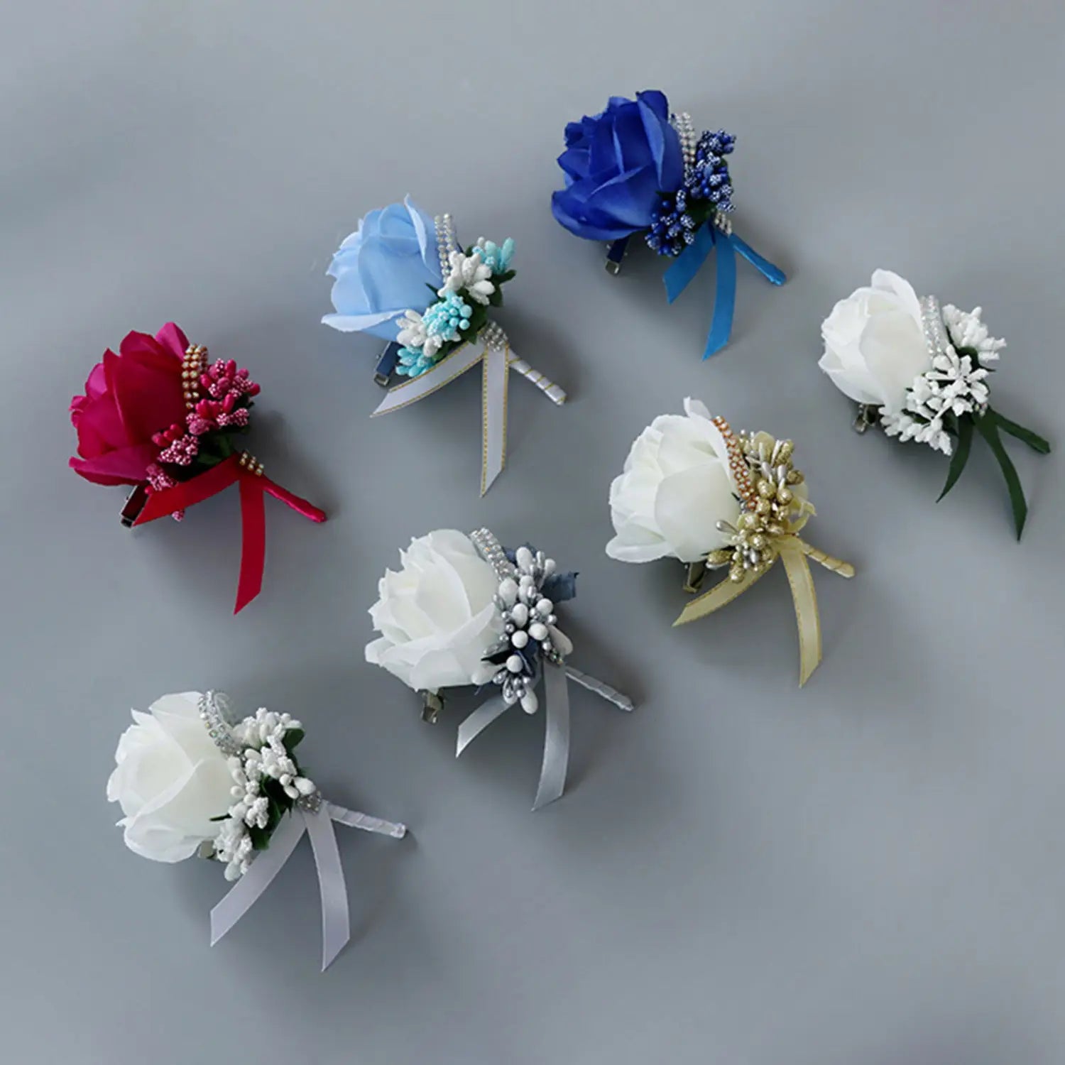 A collection of elegant artificial silk rose boutonnieres in vibrant red soft blue and pristine white adorned with delicate beads and ribbons designed for weddings and formal occasions.