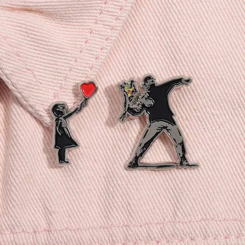 Deux épingles émaillées présentant des motifs d’art de rue emblématiques de Banksy : un enfant tenant un ballon en forme de cœur rouge et une silhouette dynamique dans une pose, reposant sur un tissu rose pâle.