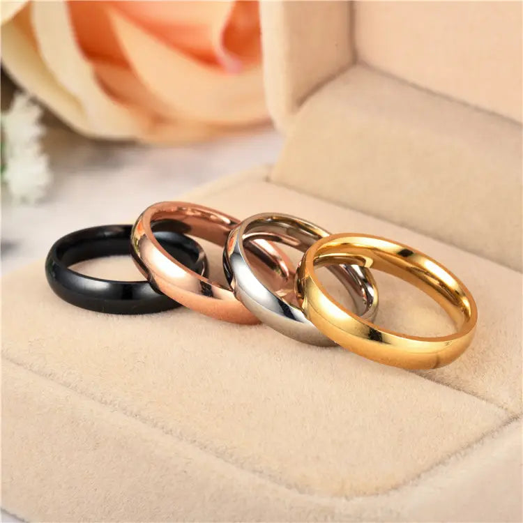Four sleek polished stainless steel wedding bands in black, rose gold, silver, and yellow gold displayed on a soft beige cushion with a 6mm rose gold ring featured prominently.