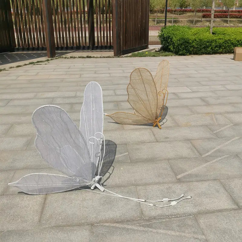 Zwei Schmetterlingsflügel-Requisiten zum Tragen in den Farben Weiß und Beige mit silbernem Mesh-Design-Trageriemen, dargestellt in einer leuchtenden, an der Decke hängenden Hochzeitstennenlichtanlage mit Schmetterlingsflügeln.