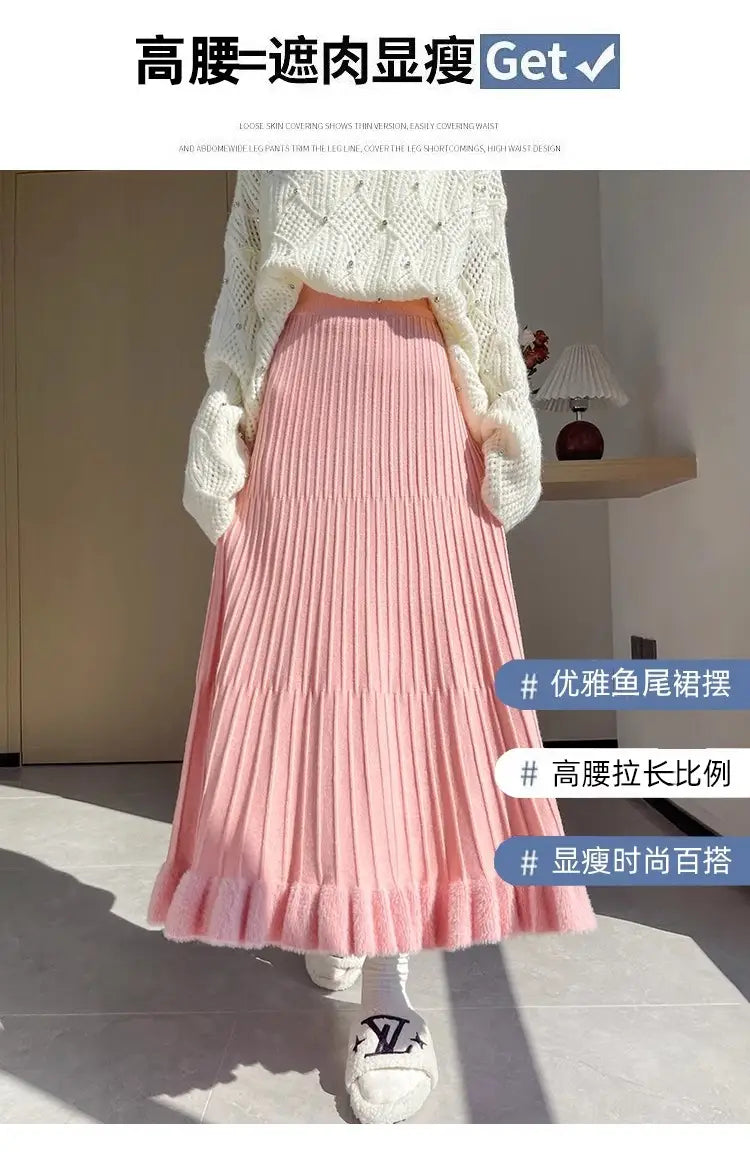 Jupe mi-longue en maille rose tendre, avec plis à taille haute et ourlet évasé, agrémentée d’une bordure volante délicate.